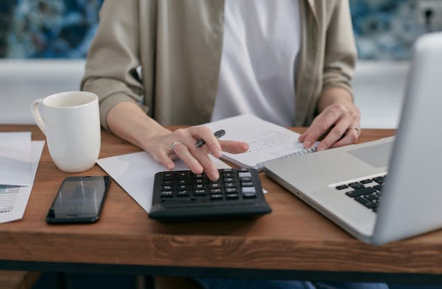 A person calculates using a calculator at a wooden desk, surrounded by papers, a smartphone, a mug, and a laptop.