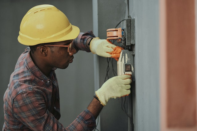 A man wearing a yellow hard hat and gloves fixes electrical wiring on a wall.