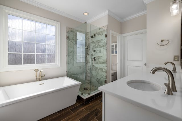 Modern bathroom with a marble-accented glass shower, sleek white bathtub under a large window, and a double sink vanity on dark wood floors.